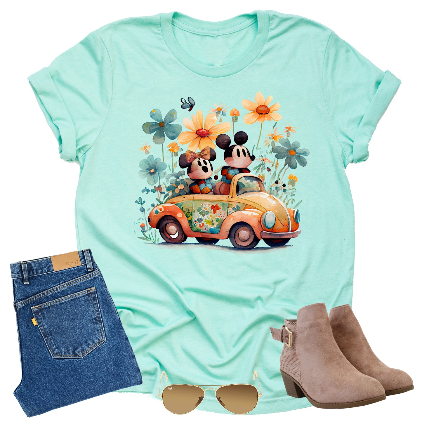 Mint green t-shirt with cartoon design of mickey and minnie characters in a car surrounded by flowers, paired with jeans, boots, and sunglasses on a white background.